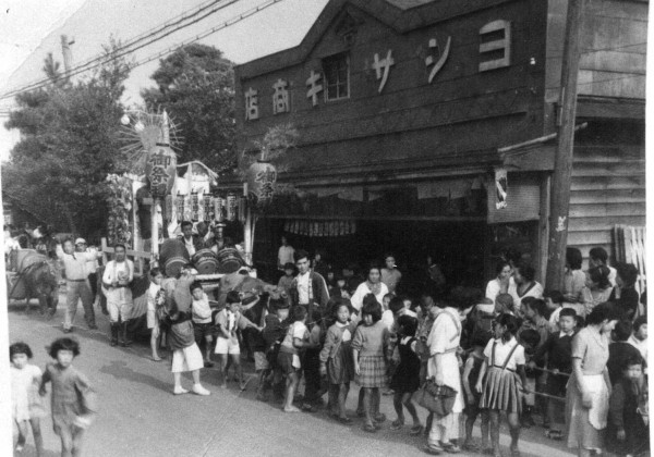 ヨシザキ商店の前で、山車を引く数十人の大人や子どもが写っている