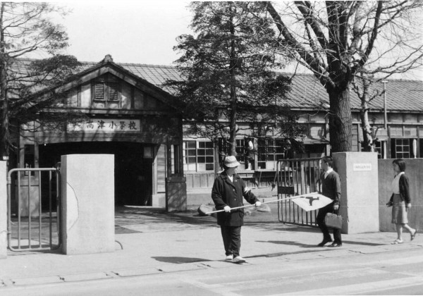 高津小学校の木造校舎と校門の写真。学童擁護員の女性が旗を手にして中央に立っている